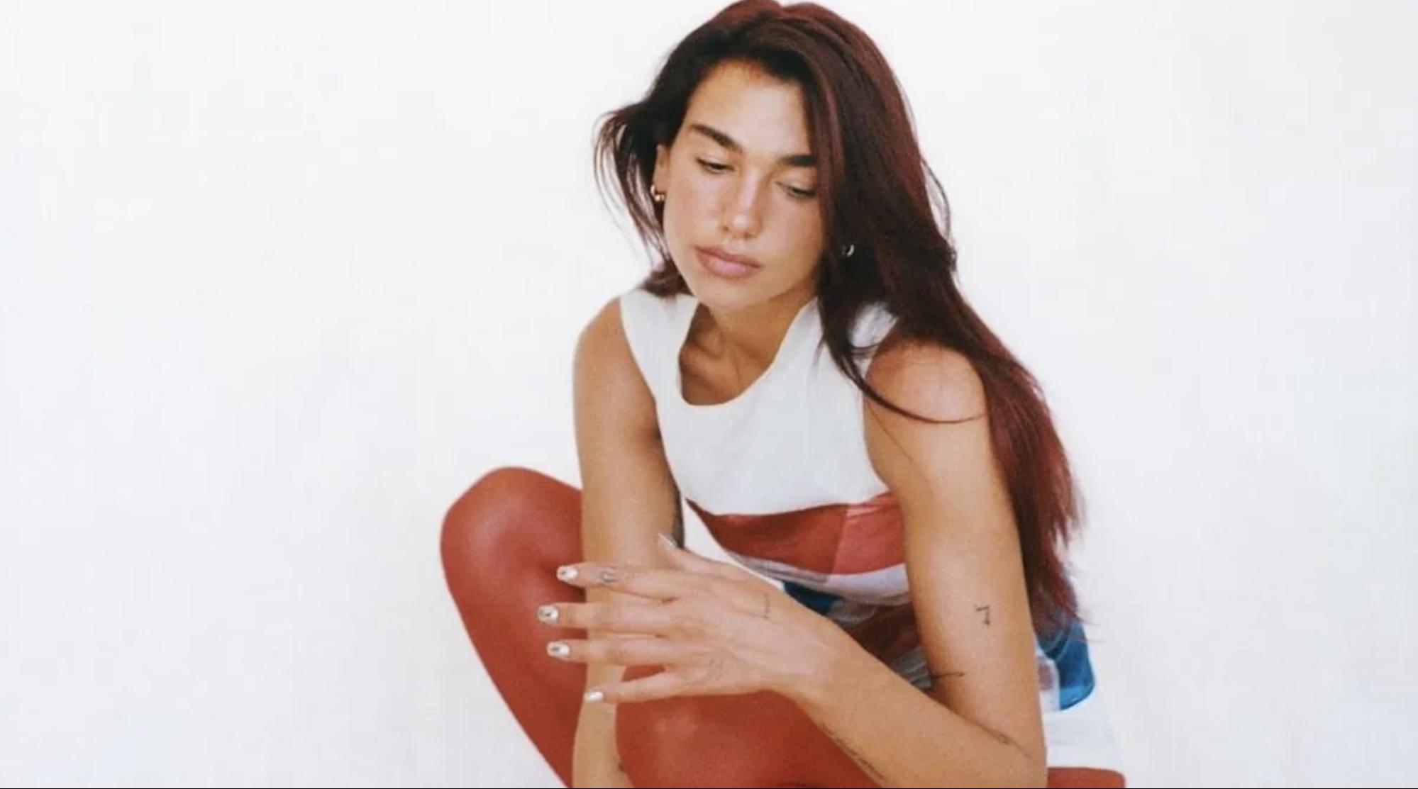 Foto da cantora Dua Lipa, mulher branca de cabelos ruivos. Ela está agachada e olhando para baixo. A artista veste uma blusa regata branca com uma linha vermelha e utiliza uma meia-calça vermelha.
