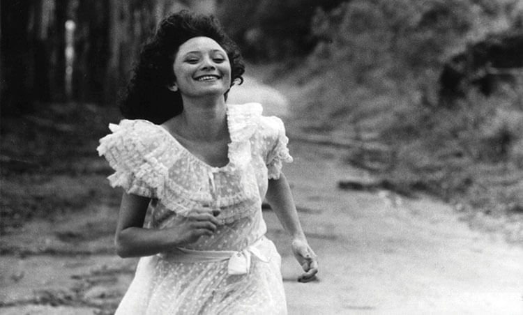 Foto da atriz Marcélia Cartaxo, uma mulher branca de cabelos cacheados e castanhos. Ela está correndo e usa um vestido branco. A foto está em preto e branco.