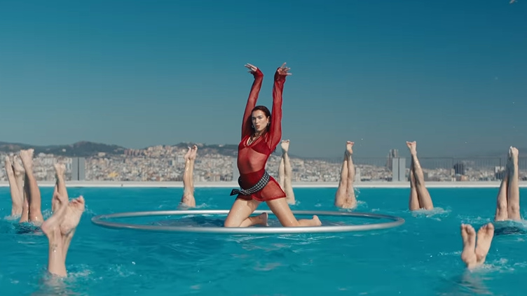 Foto da cantora Dua Lipa, mulher branca de cabelos ruivos. Ela está dentro de um círculo prata ao redor de seus dançarinos, os quais estão mergulhando na água.