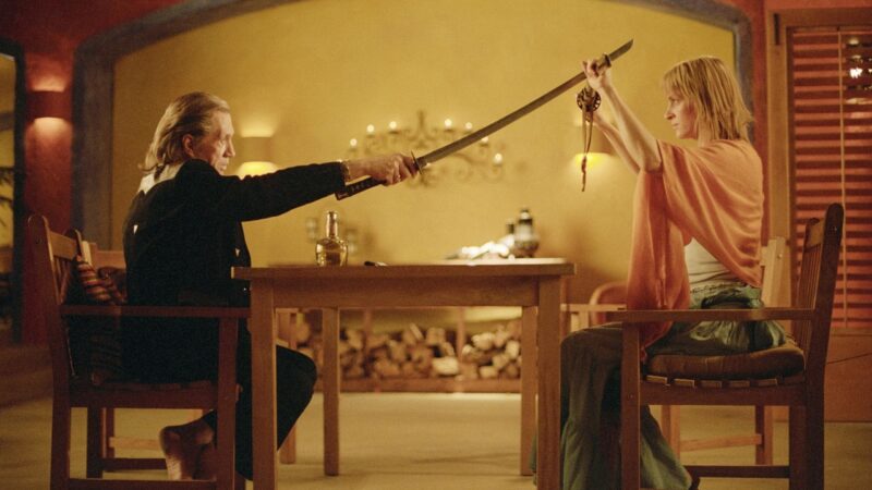 Cena do filme Kill Bill - Volume 2. Na imagem, vemos Bill vestido com uma roupa preta e atacando Beatrix com uma espada. Beatrix se defende usando a própria espada. Ela está vestida com uma calça verde e uma blusa laranja. Ambos estão sentados em uma cadeira e separados por uma mesa.