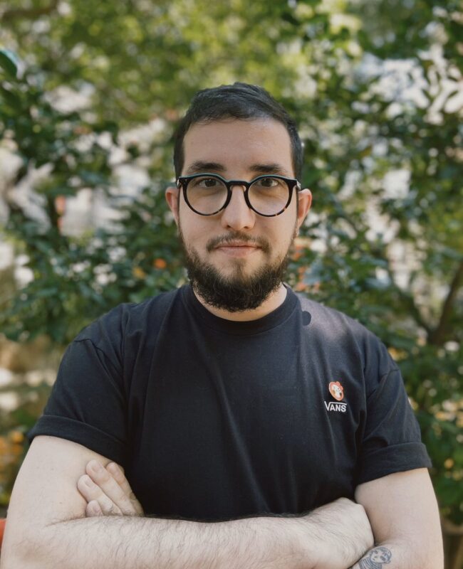 Foto do autor Vitor Martins. Vitor é um homem branco de cabelos curtos castanhos e olhos também castanhos. Ele veste uma camiseta preta e está em uma pose com os dois braços cruzados, também usa um óculos preto de armação arrendondada. Ao fundo, é possível ver a copa de uma árvore.