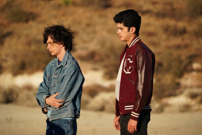 A imagem mostra dois adolescentes, Aristóteles e Dante, de pé em um deserto. Aristóteles veste uma jaqueta vermelha com letras brancas e parece sério, enquanto Dante, vestindo jeans, com os cabelos bagunçados e um hematoma no rosto, cruza os braços e olha para baixo.