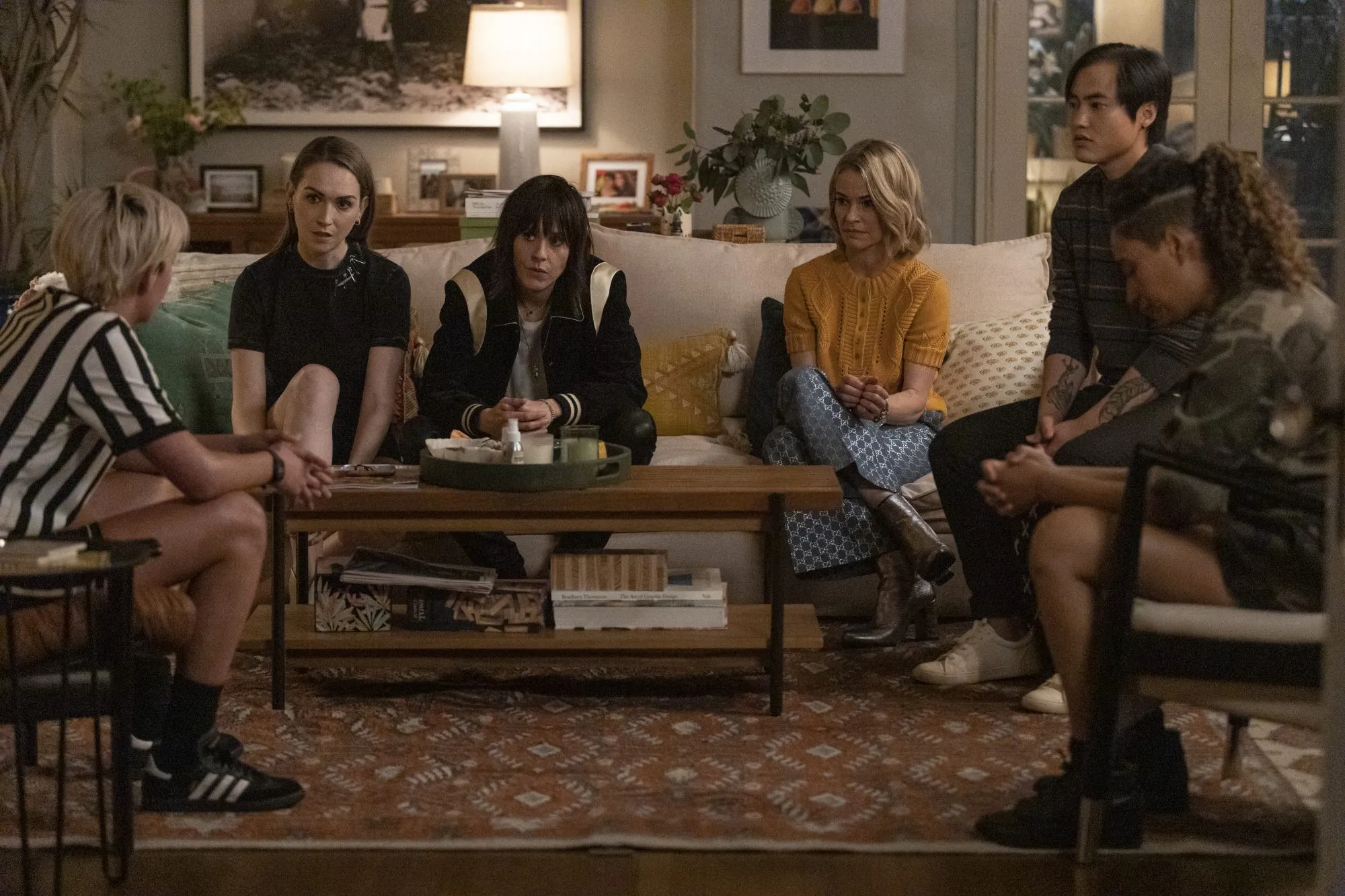 Cena de The L Word: Generation Q. Na imagem vemos parte do elenco da série reunido e se abraçando pelas laterais. Da esquerda para a direita encontra-se Finley, Shane, Alice, Bette, Dani, Sophia e Micah, respectivamente. Finley, mulher branca de cabelos curtos, usa calça marrom clara e camiseta listrada preta, vermelha e verde. Shane, mulher branca de cabelos castanhos curtos, veste uma regata branca e jaqueta de couro preta, em seu pescoço carrega um colar. Alice, mulher branca com cabelos loiros, usa um macacão jeans claro. Bette, mulher biracial de cabelos castanhos cacheados, veste uma calça marrom e uma camiseta social na cor branca que encontra-se com alguns botões desabotoados. Dani, mulher branca de cabelos castanhos escuros compridos, usa um cropped branco com uma amarração na frente com listras pretas e calça preta. Sophia, mulher negra de cabelos pretos cacheados, usa uma camiseta branca florida azul e uma jaqueta preta. Micah, homem de pele branca e cabelos pretos curtos, veste uma camiseta de botão na cor roxa estampada com desenhos em formato de V. Todos estão com sorriso no rosto. Ao fundo é possível ver uma placa azul “Welcome to SilverLake Sunset Junction” escrita em letras brancas e árvores verdes com flores rosas. O céu está na cor amarela e azul claro.