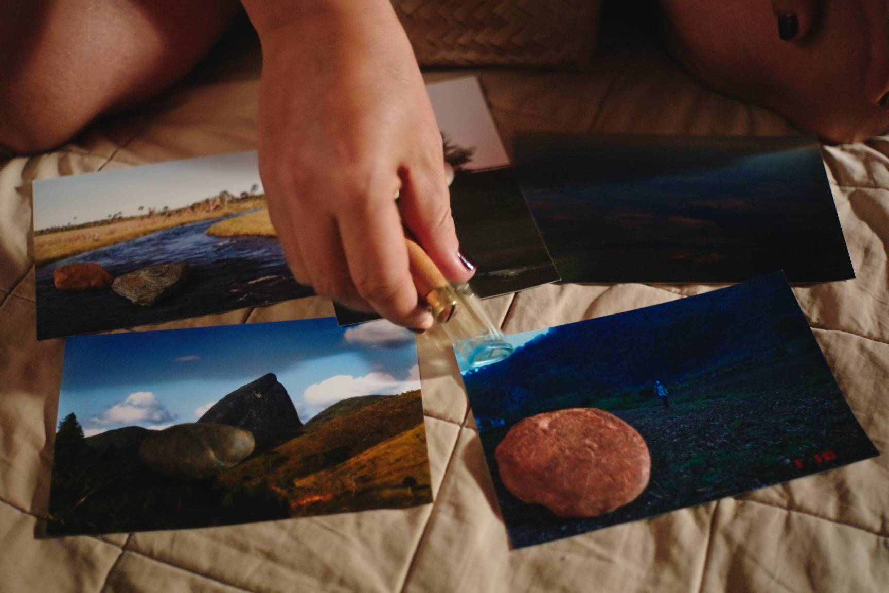 Cena do filme O Estranho. Quatro fotos estão sobre a cama. Em cada foto, há pelo menos uma pedra de diferentes formatos, cores e materiais. Uma mão está no centro da imagem colocando um objeto sobre as fotos.