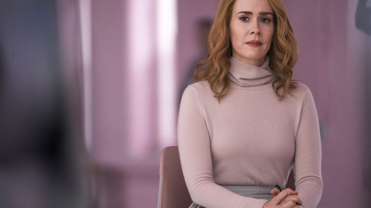 Cena do filme VidroNa imagem, a personagem da atriz Sarah Paulson está sentada com expressão de preocupação. Ela está vestindo uma blusa de gola alta e mangas compridas da cor rosa claro. Ela está com as mãos na frente do corpo, com os dedos entrelaçados apoiados na coxa. Ela é uma mulher branca, de cabelos ruivos e olhos castanhos. Na boca ela usa um batom vermelho escuro. O fundo da cor roxa está desfocado. 