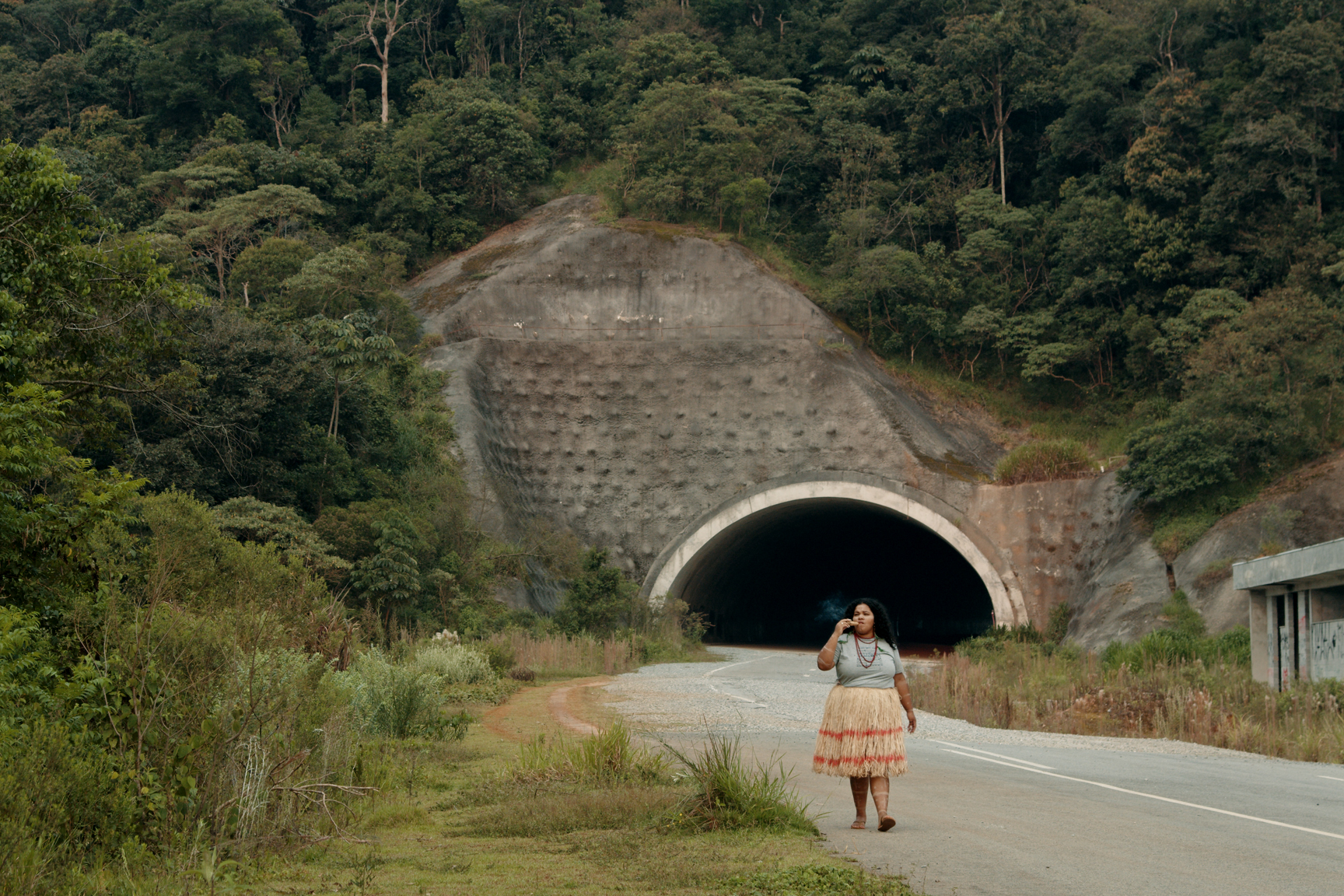 Cena de O Estranho. Ao fundo, um túnel em volta de uma floresta. Em primeiro plano, uma mulher indígena, do povo Pankararu, caminha pela estrada fumando um cachimbo. Ela veste uma blusa cinza, uma saia de palha com listras vermelhas e utiliza colares.