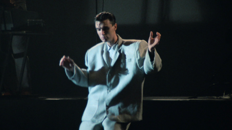 Cena de Stop Making Sense. Nela vemos David Byrne, um homem branco de cabelos pretos. Ele veste um terno duas vezes maior que ele, camisa cinza e calça cinza. Ele está com as duas mãos levantadas e o corpo levemente arqueado, pois está dançando. Ao fundo, um palco.