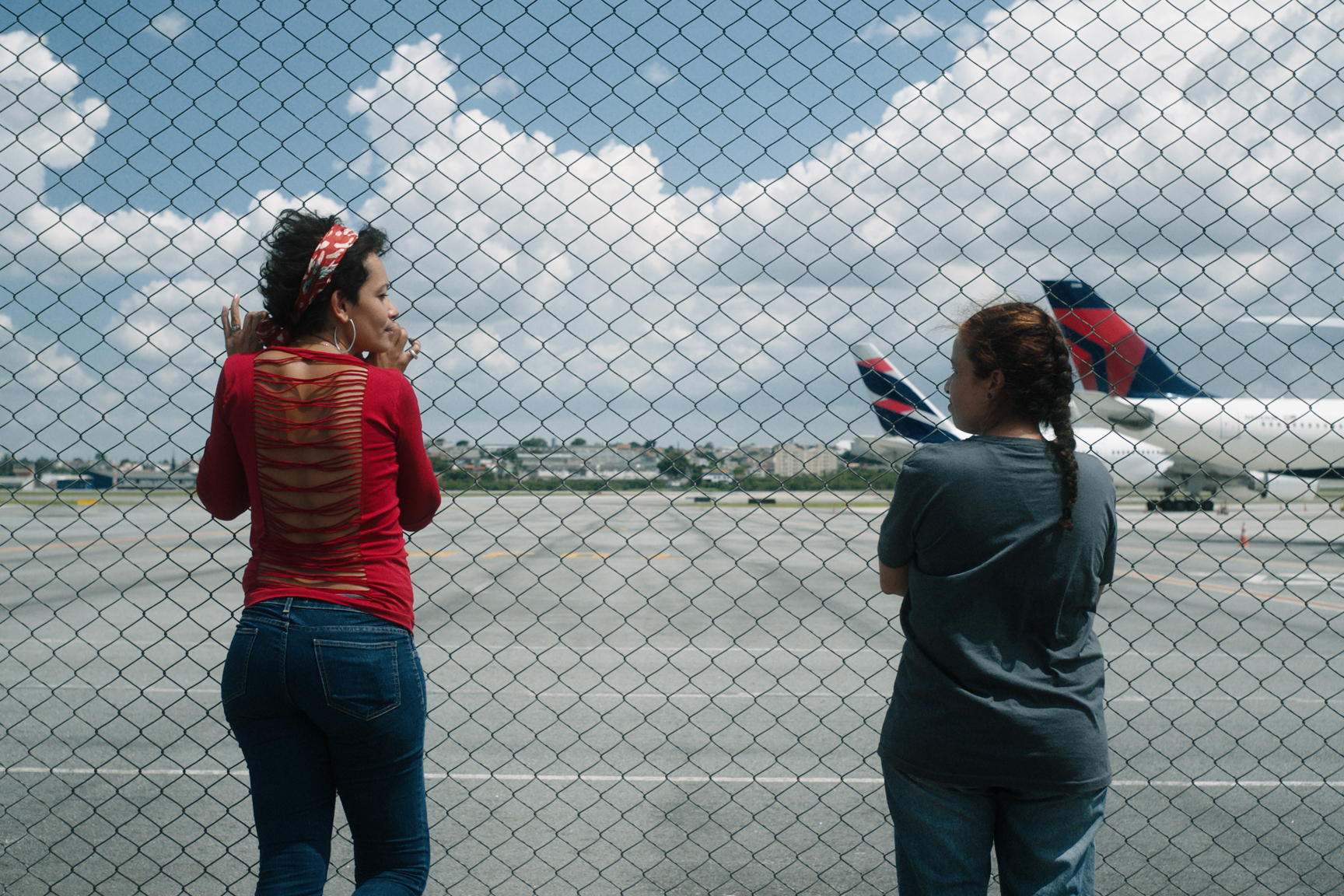 Cena do filme O Estranho. Ao fundo, vemos um avião no lado direito da imagem. Em primeiro plano, uma grade divide o aeroporto das duas mulheres que estão do lado de fora da grade. Ao lado esquerdo, Sílvia está apoiada na grade. Utiliza uma tiara colorida, uma blusa vermelha com uma abertura nas costas e calça jeans. Mais a direita, Alê está de braços cruzados. Utiliza uma camisa cinza, calça jeans e seu cabelo está amarrado com uma trança.