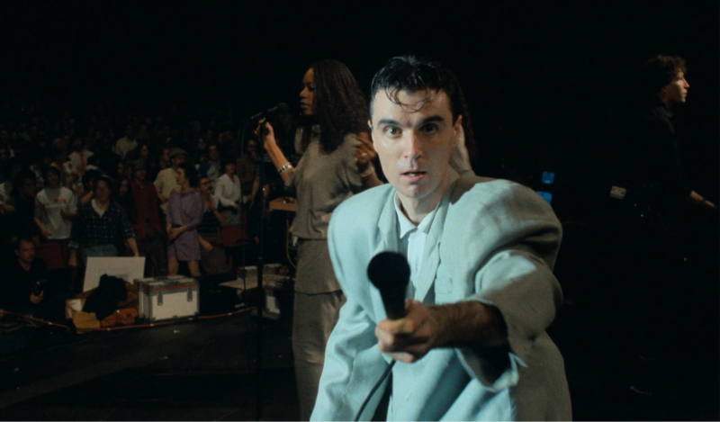 Cena de Stop Making Sense. Nela vemos David Byrne, um homem branco de cabelos pretos. Ele veste uma camisa cinza e um terno cinza, que é duas vezes maior que seu tamanho. Ele está segurando um microfone com a mão esquerda e aponta ele em direção a câmera. Ao fundo, as backing vocals da banda Talking Heads, da qual Byrne faz parte.