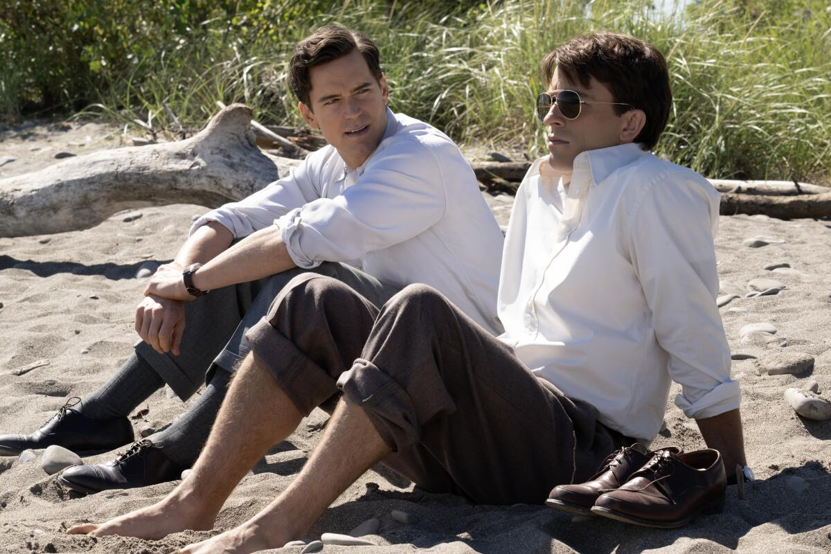 Imagem da série Companheiros de Viagem. Na foto, os atores Jonathan Bailey e Matt Bomer estão sentados na areia da praia. Os dois estão utilizando uma camisa branca de botão e calças com tons acinzentados. Jonathan Bailey está usando óculos de sol e Mat Bomer está olhando para ele.