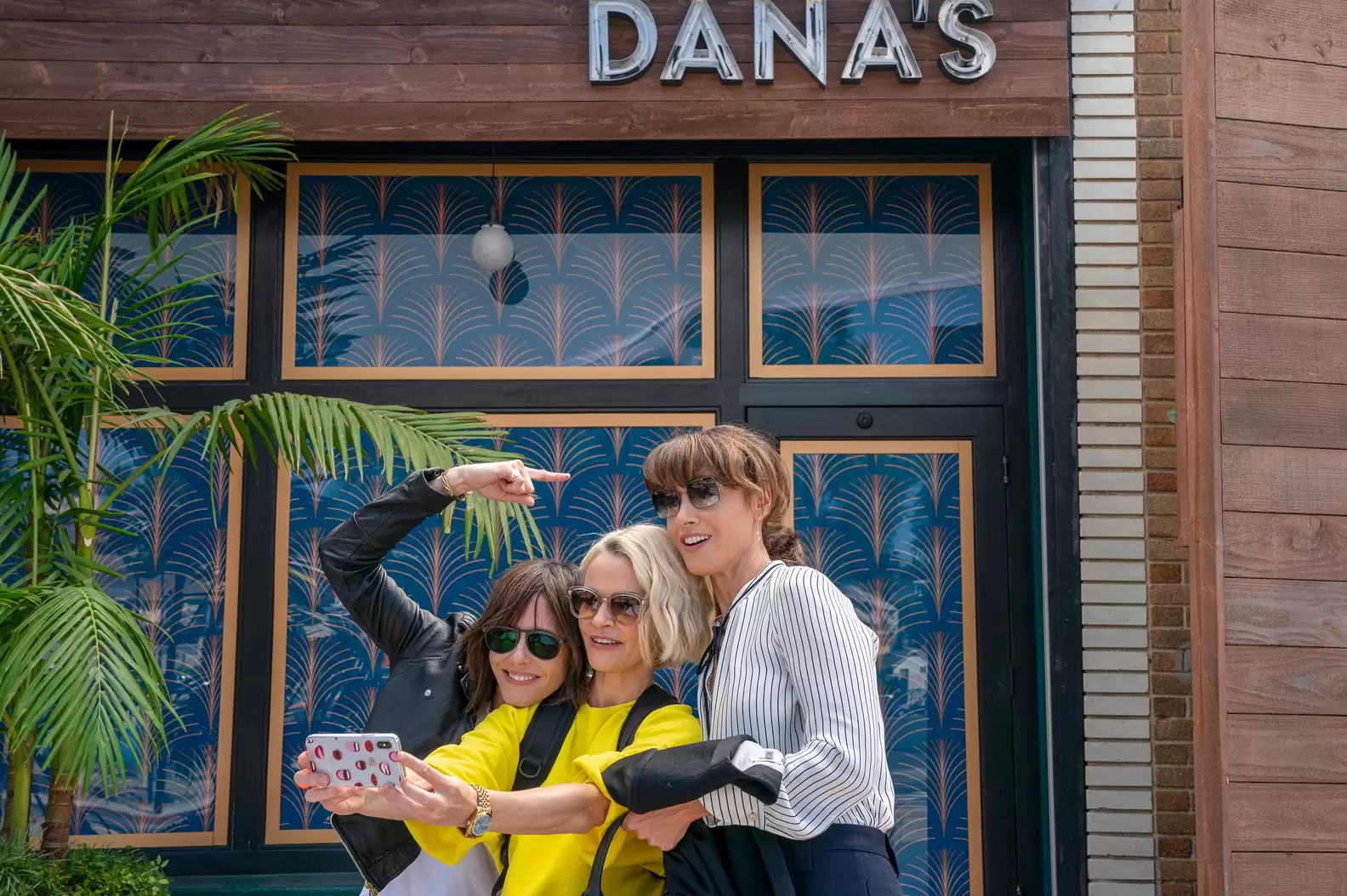 Cena da série The L Word: Generation Q. Na imagem vemos Shane, Alice e Bette tirando uma selfie na frente de um estabelecimento. Shane, mulher branca de cabelos castanhos escuros, veste uma camiseta branca e jaqueta de couro preta, seu braço esquerdo está esticado para cima de sua cabeça e seu dedo apontado para cima. Alice, mulher branca de cabelos loiros, veste uma camiseta amarela e segura um celular com suas duas mãos. Bette, mulher biracial com cabelos castanhos claros, veste uma calça social preta e uma camiseta social listrada branca e preta, em sua mão segura uma bolsa e em seu braço encontra-se um blazer, ambos na cor preta. Todas estão usando óculos de sol escuros. Centralizado ao fundo da imagem existe uma grande janela de vidro, do lado esquerdo é possível ver uma palmeira e no canto superior direito encontra-se um letreiro escrito “Dana’s” na cor branca.