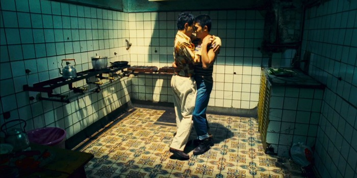 Dois homens abraçados dançam ao som de tango na cozinha de casa, em um momento íntimo. Leslie Cheung, à esquerda, veste uma camisa neutra e calças bege, enquanto Tony Leung, à direita, usa camisa neutra com calças jeans azuis.