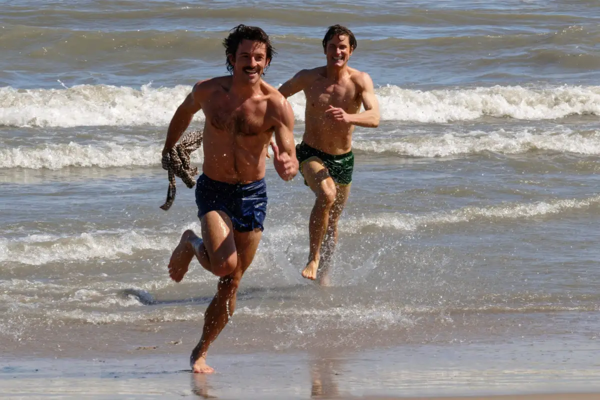 Imagem da série Companheiros de Viagem. Na foto, os atores Jonathan Bailey e Matt Bomer estão em uma praia. Os dois estão molhados e estão correndo pela areia. Eles vestem um shorts de banho.