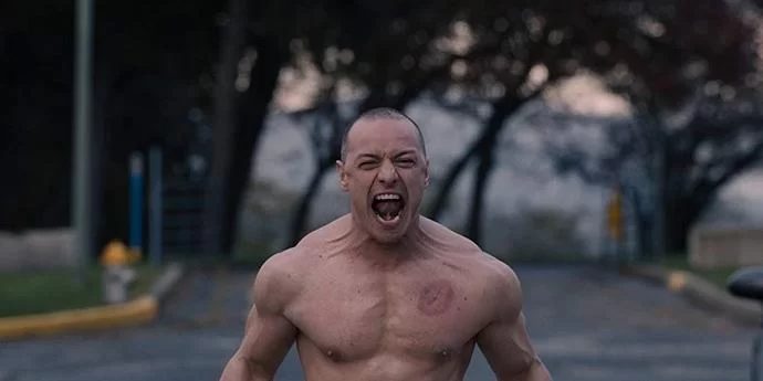 Cena do filme VidroNa imagem, o personagem Kevin está gritando com muita força, aparecendo suas veias no pescoço. Ele está sem camiseta e tem um machucado no peito esquerdo. Kevin é um homem branco na faixa dos 40 anos, careca. O fundo está desfocado, mas está de dia e há árvores. 