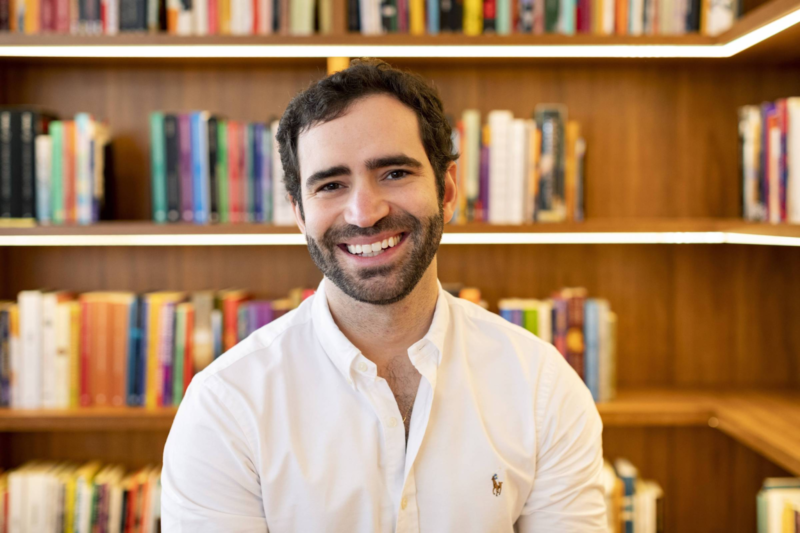 Foto de Pedro Pacífico, conhecido nas redes sociais como bookster. Homem de 30 anos, ele é branco, tem cabelos curtos pretos e barba preta.Pedro sorri e está centralizado na foto. Ele veste uma camisa social. O fundo da foto é uma estante com vários livros desfocados]