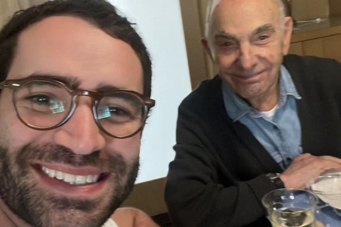 Foto de Pedro Pacífico, conhecido nas redes sociais como bookster, ao lado do padrinho. Pedro tem 30 anos, é branco e tem cabelos curtos pretos e barba preta.Pedro sorri e está ao lado do padrinho, que está no lado direito da foto. O idoso veste um casaco preto e uma blusa social azul. Ele também sorri para a foto.