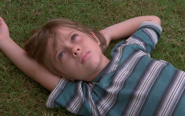 Cena do filme Boyhood: Da Infância à Juventude. Um menino branco, loiro de olhos azuis deitado na grama verde com um braço sob a cabeça. Ele veste uma camisa listrada azul, branca e cinza.