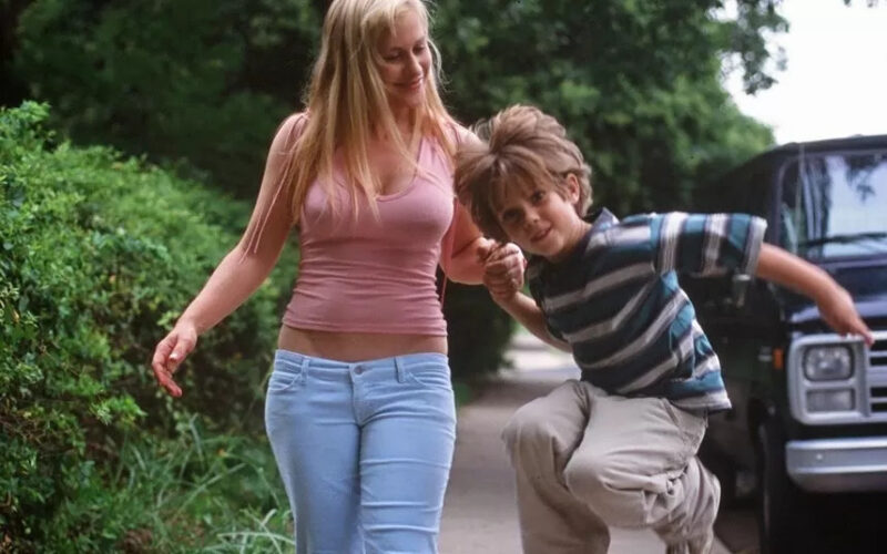 Cena do filme Boyhood: Da Infância à Juventude. No fundo um arbusto verde com um carro preto. No centro está na calçada uma mulher branca, loira, de regata rosa e calça jeans segurando a mão de um menino branco, loiro que veste uma blusa listrada nas cores verde, cinza e preto e uma calça cinza. Ele está pulando.