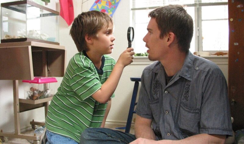 Cena do filme Boyhood: Da Infância à Juventude. Ambientando em um quarto com as paredes brancas, uma janela e alguns terrários ao fundo. No primeiro plano está Mason e o pai. Mason é uma criança branca de cabelos loiros e veste uma camisa listrada verde e branca, está observando uma lupa apontada para o rosto do seu pai. O pai está a sua frente, um homem branco de cabelos castanhos e cavanhaque, veste uma camisa jeans com detalhes de costura.