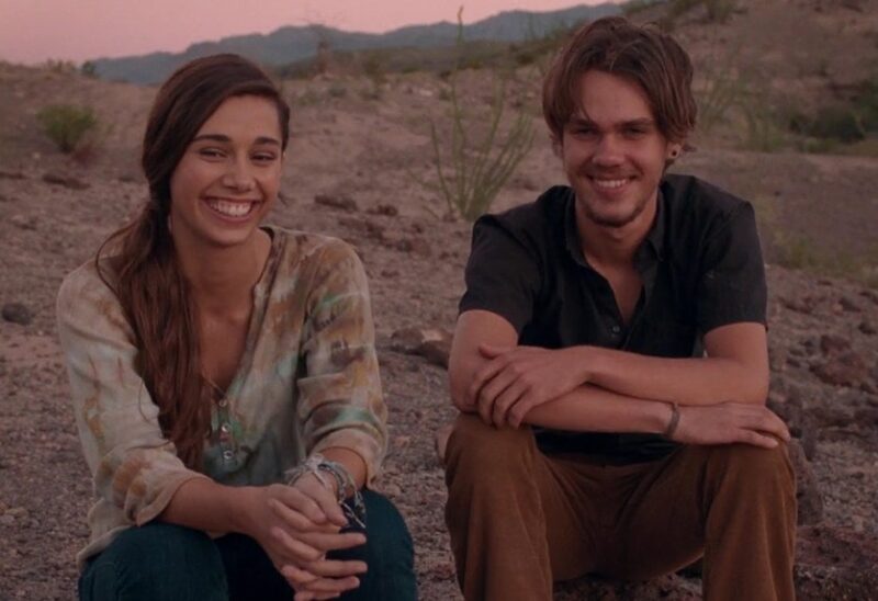 Cena do filme Boyhood: Da Infância à Juventude. Ambientando em uma montanha com o pôr do sol ao fundo. A direita está Mason, um homem branco de cabelos compridos divididos ao meio, com barba e bigode, ele usa brinco na orelha direita e veste uma camisa preta e calça marrom. A esquerda está uma garota branca de cabelos castanhos amarrados de lado, veste uma camisa estampada e calça jeans. Ambos estão sentados e sorrindo.