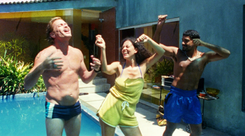 Cena do filme Motel Destino. Da esquerda para a direita: Elias, Dayana e Heraldo. Os homens estão de sunga e shorts e a mulher com um conjunto curto verde. Eles estão dançando no sol na frente de uma piscina.