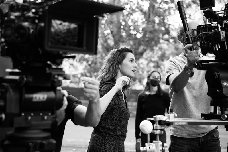 Cena da produção do filme Ainda Temos o Amanhã. Em preto e branco está a foto da diretora e atriz principal Paola Cortellesi, uma mulher branca, italiana de cabelos ondulados na altura dos ombros. Ela está encarando uma câmera ao ar livre enquanto um cinegrafista a seu lado a manuseia. Em primeiro plano temos uma câmera desfocada à esquerda que ocupa metade da imagem e ao fundo uma paisagem ao ar livre com uma mulher usando máscara.