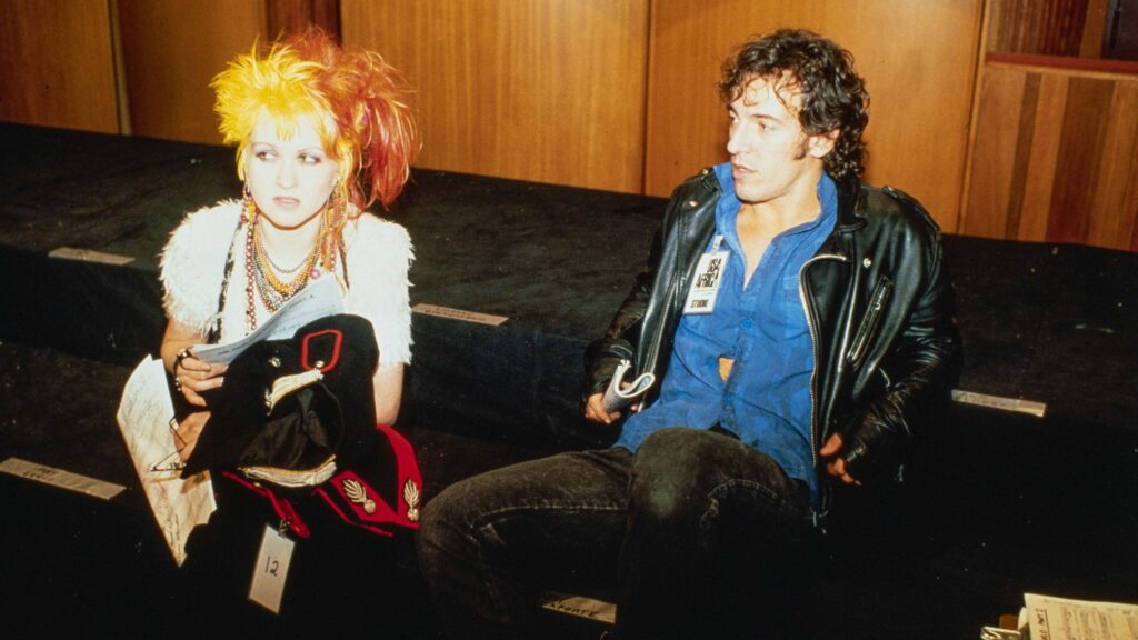 Cena de A Noite que Mudou o Pop. Da esquerda para a direita na imagem, Cyndi Lauper e Bruce Springsteen estão sentados no estúdio de gravação. Lauper, uma mulher branca de cabelos loiros e coloridos, veste uma camiseta branca e muitos acessórios enquanto segura um papel com os versos de We Are The World. Já Springsteen, um homem branco de cabelos escuros, veste jaqueta de couro preta sobre uma blusa azul enquanto olha para a cantora. Ao fundo, o cenário apresenta tons amarronzados.