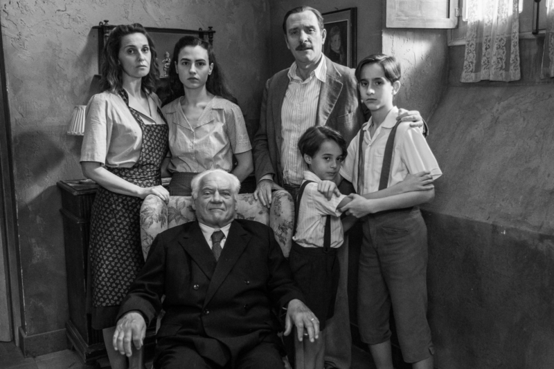 Cena do filme Ainda Temos o Amanhã. Em preto e branco está uma família de seis pessoas posicionadas para foto. À direita está Délia, uma mulher branca, italiana de meia idade com cabelos ondulados escuros na altura do ombro, vestindo um avental e camiseta. Ao seu lado esquerdo está Marcella, sua filha branca de cabelos longos pretos e camiseta polo. Abaixo está o Avô Ottorino que usa um terno preto e está sentado em uma poltrona. Ao seu lado estão dois garotos com camisetas e suspensórios abraçados com seu pai Ivano, um homem com um terno claro, bigode expressivo e camiseta social por baixo. Todos encaram sua frente.