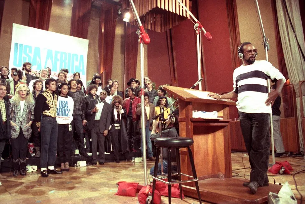 Cena de A Noite que Mudou o Pop. Na imagem, parte dos 45 artistas convocados para cantar aparecem posicionados em um estúdio de gravação. Em frente a todos eles está Quincy Jones, um homem negro, produtor da música We Are The World. Ele veste roupas casuais e usa um fone de ouvido. O cenário é composto por tons amarronzados, espumas nas paredes e um telão ao fundo dos cantores.