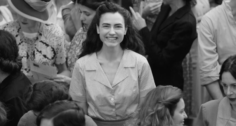 Cena do filme Ainda Temos o Amanhã. Em preto e branco está Marcella, interpretada pela atriz Romana Vergano. Uma mulher italiana, branca e jovem com cabelos longos, escuros e ondulados que sorri para a câmera. Ela está rodeada de mulheres em uma multidão e veste uma camiseta clara quadriculada polo.