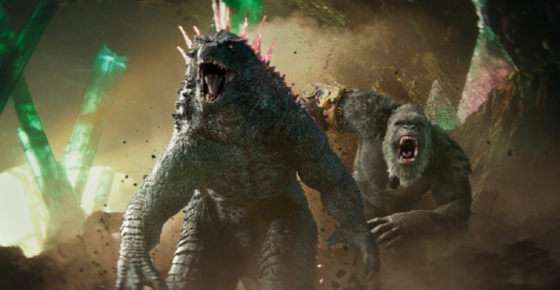 Cena do filme Godzilla e Kong: O Novo Império. A imagem mostra os dois monstros que dão nome ao filme. Do lado esquerdo, Godzilla é um monstro parecido com um dinossauro, bípede de cor cinza esverdeada, com espinhos rosas em suas costas, vai correndo na frente. Ao lado direito está Kong, um gorila gigante, que possui uma luva metálica amarela. Na cena, os dois personagens principais do filme estão correndo em direção a tela. Godzilla vai correndo na frente. Logo atrás dele vem Kong,de boca aberta, preparado para o ataque. No plano de fundo, um cenário rochoso destruído com o que parece ser cristais verdes.