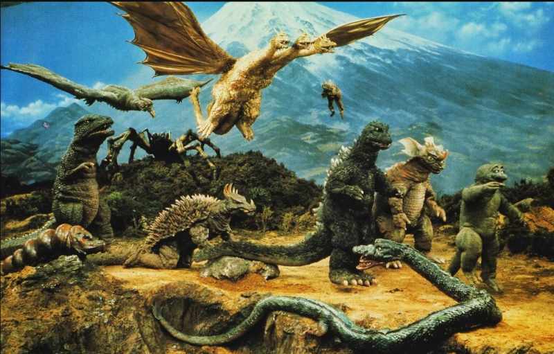 Cena do filme O Despertar dos Monstros. É um cenário de floresta com o monte Fuji ao fundo. O plano principal contém diversas pessoas fantasiadas interpretando monstros gigantes. A maioria deles se parecem com dinossauros, com Godzilla sendo o mais chamativo da imagem. Mas também temos criaturas que se assemelham a dragões,cobras, aranhas e vermes gigantes.