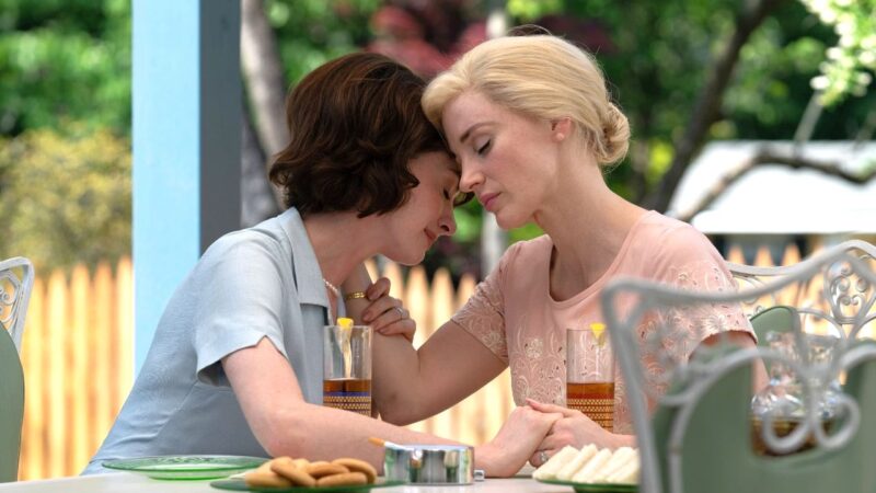 Cena do filme Instinto Materno. As personagens Céline e Alice interpretadas, respectivamente, por Anne Hathaway e Jessica Chastain, estão sentadas à mesa em uma varanda externa. Céline é uma mulher branca com cabelos castanhos e usa um vestido azul-claro típico dos anos 1960. Alice é uma mulher branca com cabelos muito louros e usa um vestido rosa. As duas estão com as mãos dadas.