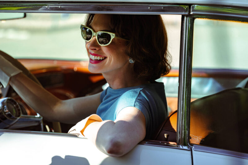 Cena do filme Instinto Materno. A personagem Céline, interpretada por Anne Hathaway, sorri dentro de um carro antigo. A personagem tem pele branca e apoia um braço na porta do carro, ao passo que a sua outra mão está segurando o volante. Celine usa óculos escuros e tem um cabelo castanho curto.