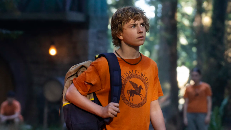 Cena de Percy Jackson e os Olimpianos. Nela vemos Percy, um adolescente branco de cabelos loiros. Ele vesta uma camiseta laranja com uma estampa de um pégaso com os dizeres "CAMP HALF BLOOD" estilizado em letras gregas, e uma mochila em somente um dos ombros. Ele está olhando para cima e, ao fundo, há uma floresta