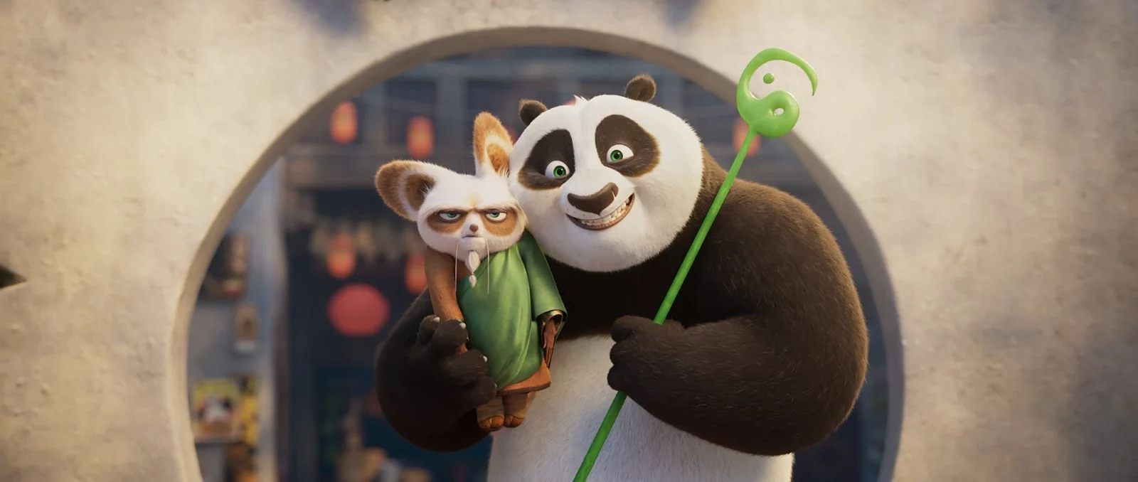 Cena do filme Kung Fu Panda 4. Po (um panda) segura Mestre Shifu (um coala vermelho) e o abraça encostando suas cabeças uma na outra. Po sorri e Shifu mantém um semblante irritado enquanto olham para a frente; Shifu veste sua túnica verde e laranja. Eles estão parados em frente à entrada do restaurante de um dos pais de Po, uma parede de pedra cinza clara com uma abertura circular no centro; o panda segura o cajado verde da sabedoria dado a ele por mestre Oogway.