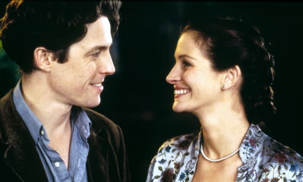 Cena do filme Um Lugar Chamado Notting Hill. À esquerda está Hugh Grant, um homem branco com cabelos castanhos que veste uma camisa azul e um blazer marrom. À direita, vemos Julia Roberts, uma mulher branca com cabelos castanhos trançados. Ela veste uma camisa branca com vários detalhes florais e um colar de prata. A imagem mostra os perfis dos dois atores, ao passo que eles se olham e sorriem.