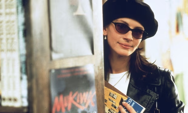 Cena do filme Um Lugar Chamado Notting Hill. Na imagem vemos Julia Roberts, uma mulher branca com cabelos castanhos. Ela veste uma camiseta branca, uma jaqueta de couro, uma boina preta e óculos de sol. A atriz está dentro de uma livraria, e ela está atrás de uma prateleira, que está desfocada. Ela segura um livro em suas mãos.