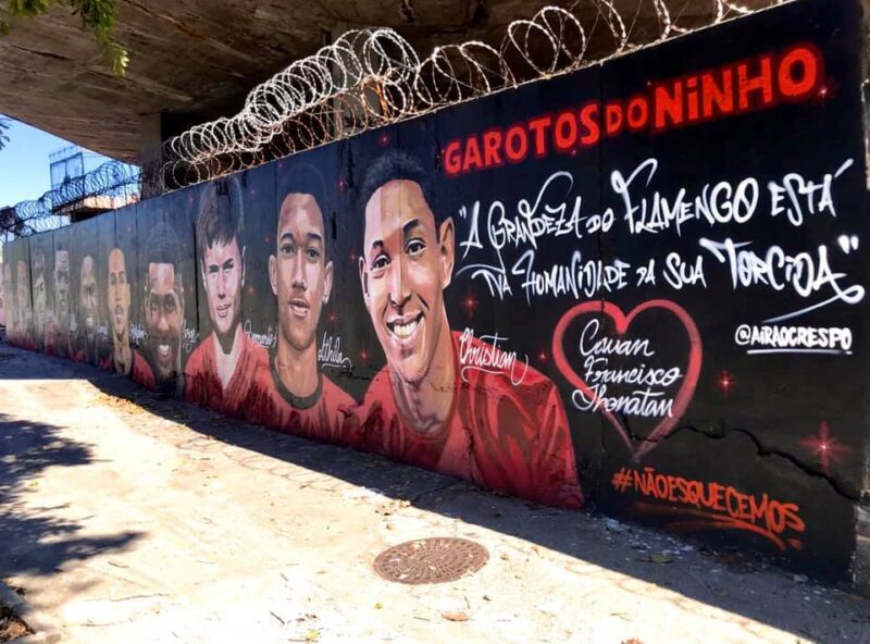  Foto do Mural do Garotos do Ninho. Tirada em um dia ensolarado, a foto retrata uma parte da homenagem, onde aparece alguns rostos e seus respectivos nomes do lado: Rykelmo, Jorge Eduardo, Bernardo, Athila e Christian. No fundo preto, ao lado da pintura de Christian, há os escritos: logo acima, em vermelho, “Garotos do Ninho”. Abaixo, em branco, a frase “A grandeza do Flamengo está na humanidade da sua torcida.”. Em baixo da frase, há um coração com os nomes, em branco, dos feridos sobreviventes da tragédia: Cauan, Francisco e Jhonatan. Do lado do coração vermelho, encontra-se a identificação, em branco, da rede social do artista Airá OCrespo (@airaocrespo). Abaixo do coração, há a hashtag em vermelho #NaoEsquecemos