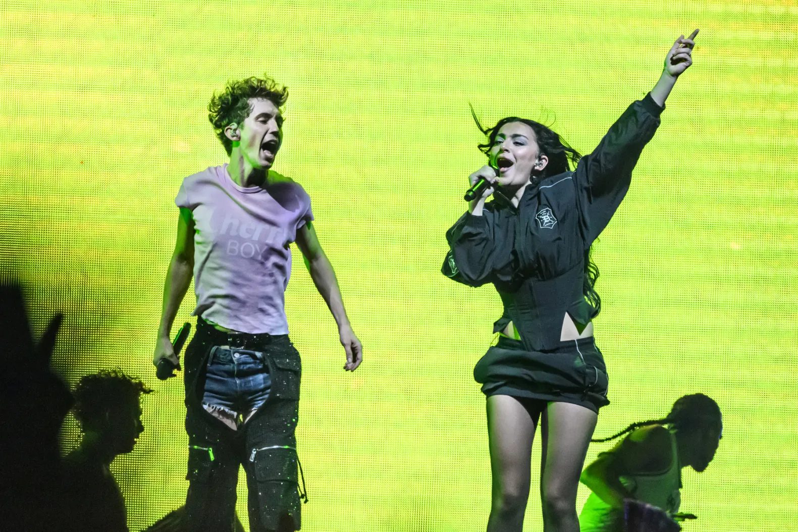 Cena da turnê Sweat Tour. Nela está o cantor australiano Troye Sivan, um homem branco de 30 anos e cabelos loiros. Ele está com uma calça aberta no centro e um short jeans por baixo, e uma camiseta branca. Ele segura um microfone e pula com a boca aberta. Ao seu lado direito está a cantora Charli xcx, uma mulher branca com cabelos pretos ondulados. Ela está cantando com seu microfone na boca e outra mão levantada, vestindo uma saia, jaqueta e meia calça pretas.