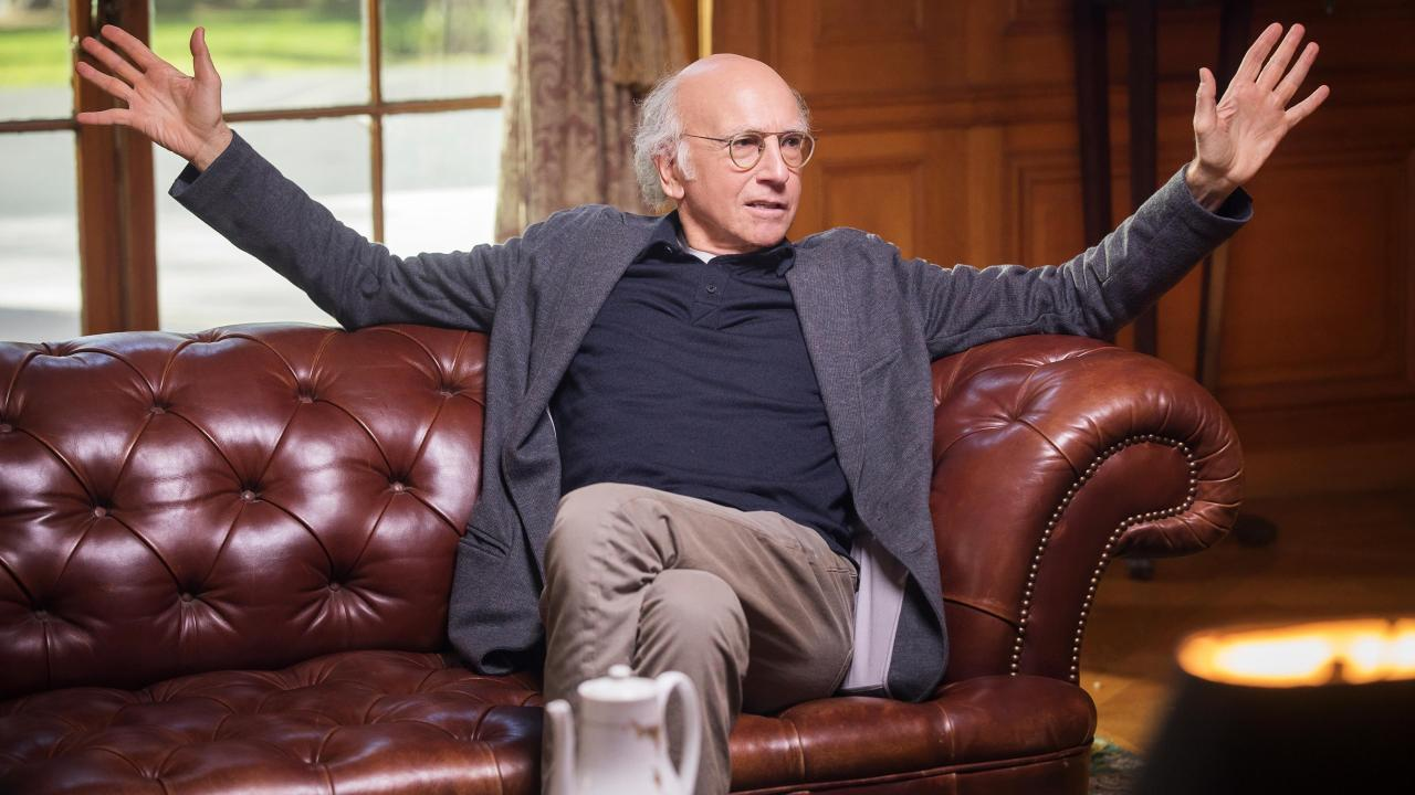 Cena da série Segura a Onda. Larry David está sentado em um sofá vinho, encostado no braço do sofá e com as pernas cruzadas. Ele está com os braços e as mãos abertas em movimento de expressão, enquanto olha para o seu lado esquerdo.