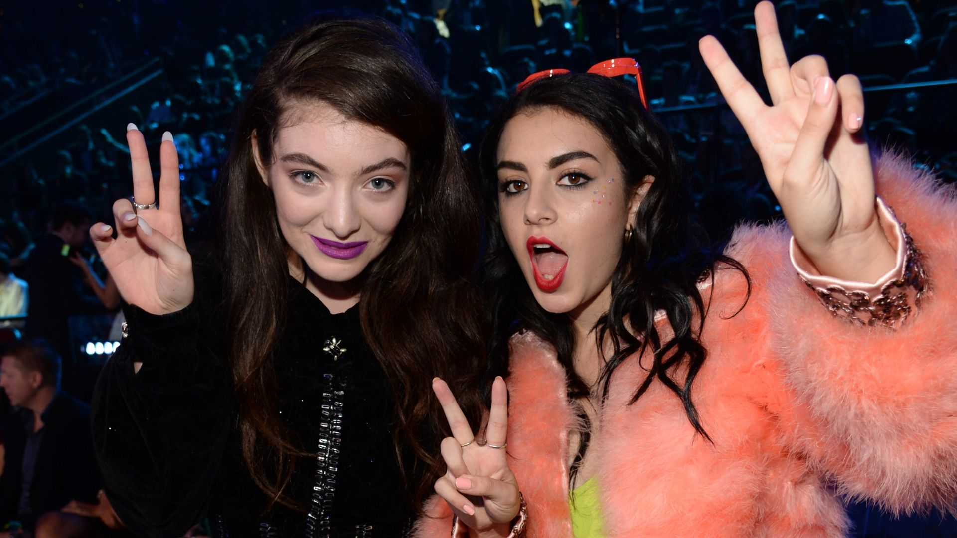 Foto das cantoras Charli xcx e Lorde em meados de 2013. Elas estão fazendo sinal de paz com seus dedos indicador e do meio. Elas são mulheres brancas de cabelos escuros ondulados. Charli está com um batom vermelho, e uma jaqueta de cor salmão felpuda. Lorde está com um batom roxo e roupas completamente pretas com detalhes em prata na gola. Elas estão em um evento noturno rodeadas de pessoas.