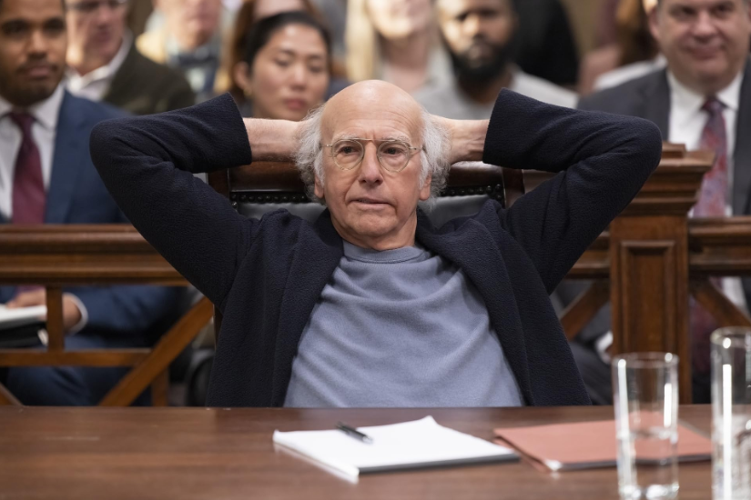 Cena da série Segura a Onda. O protagonista Larry David é um homem branco, calvo, com cabelos brancos nos lados da cabeça e óculos. Ele está sentado em uma mesa de tribunal, de costas para as pessoas sentadas nos bancos atrás dele. Ele está com os braços levantados e as mãos atrás da cabeça