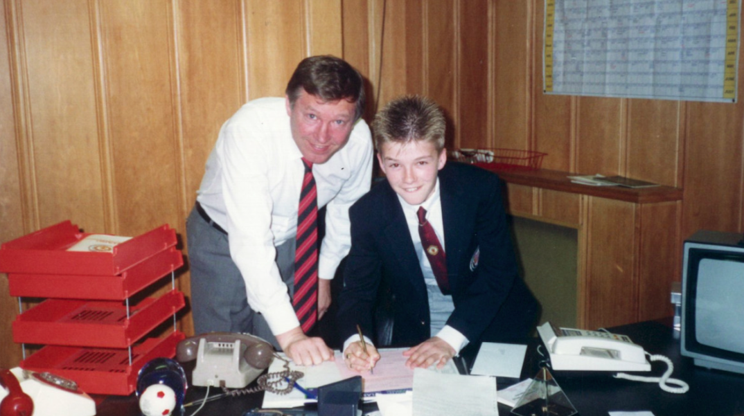 Fotografia de Alex Ferguson e David Beckham veiculada no documentário Beckham. Da esquerda para a direita na imagem, Ferguson e Beckham sorriem para a câmera que, por sua vez, os capturam a partir das cinturas. Ambos são homens brancos de cabelos e olhos claros. Alex Ferguson veste um uma camiseta branca, gravata de estampa vermelha e preta, e uma calça cinza de linho. David Beckham veste um terno preto sobre uma camiseta branca e uma gravata de estampa vermelha. Eles estão assinando um contrato em uma mesa repleta de papelada. Ao fundo, o cenário é uma sala de reuniões.