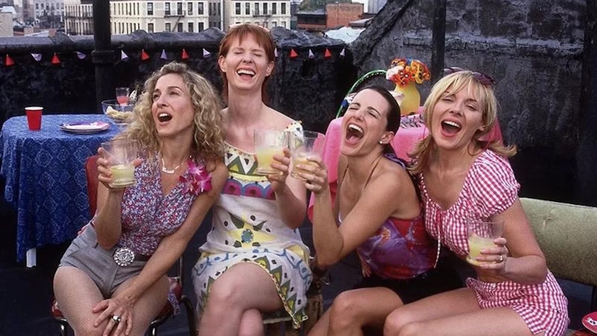 Na imagem, da esquerda para a direita, estão as personagens Carrie, Miranda, Charlotte e Samantha. Elas estão sentadas em um terraço, sorrindo e segurando copos de bebida. Carrie usa uma blusa estampada e shorts cinza, Miranda veste um vestido branco com estampas coloridas, Charlotte está com um top rosa e Samantha usa uma blusa xadrez vermelha e branca. Ao fundo, há uma mesa com toalha azul e um copo vermelho, além de um arranjo de flores.