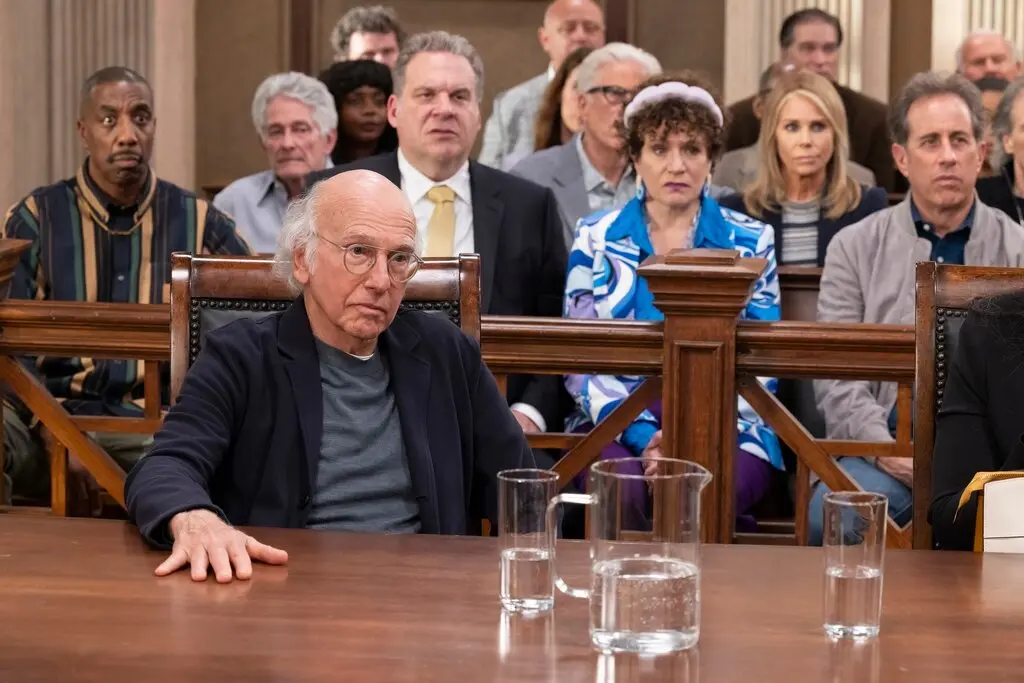 Cena da série Segura a Onda. Larry David está em uma mesa de tribunal. Nos bancos atrás dele, pode-se ver outros personagens da série sentados. Da esquerda para a direita no primeiro banco: Leon, Jeff, Susie e Jerry Seinfeld. No banco de trás, estão Ted Danson e Cheryl. Figurantes ocupam os demais lugares.