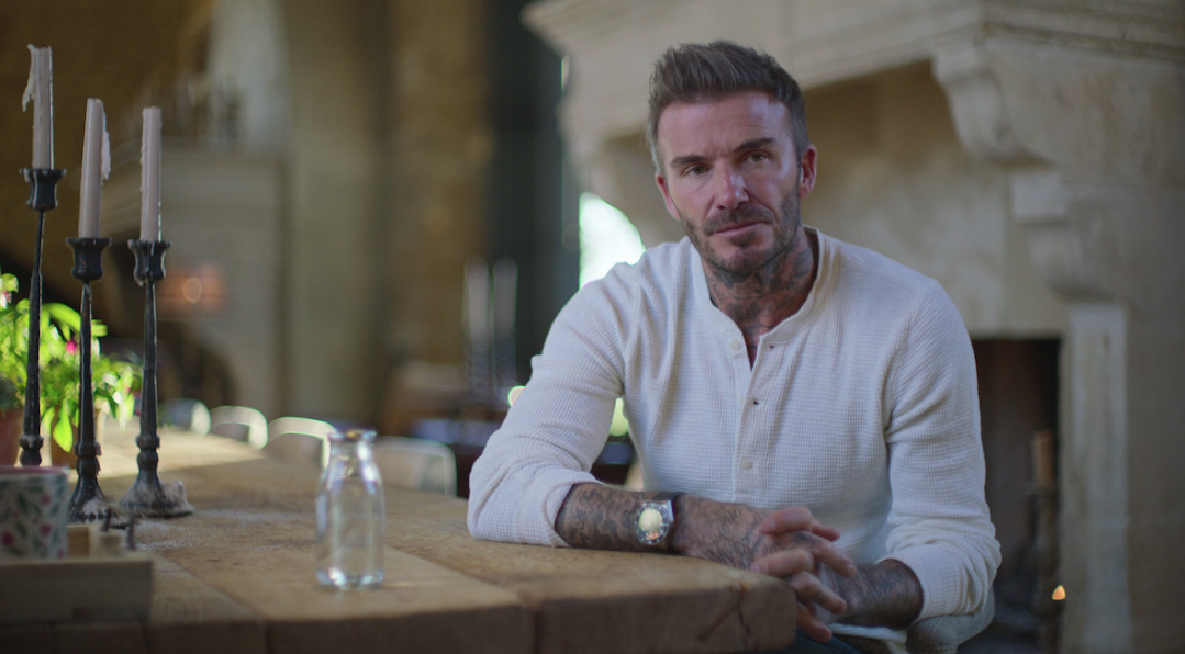 Cena do documentário Beckham. Na imagem, David Beckham aparece olhando para algo atrás da câmera que, por sua vez, o captura a partir do busto. Beckham é um homem branco de cabelos e olhos claros. Ele veste uma camiseta de algodão branca de mangas longas e um relógio preto no pulso. Ele apoia um dos braços em uma mesa de madeira, em que também estão um candelabro e uma taça transparente. Ao fundo, desfocado, é possível notar que o cenário se trata da sala aconchegante da mansão da família Beckham. Raios de sol invadem a paisagem composta por uma lareira.