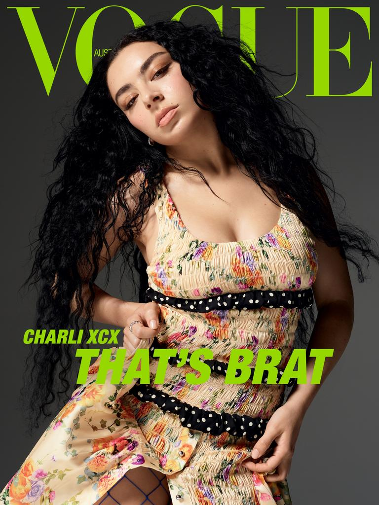 Capa da revista Vogue da australia. Ao topo está o nome da revista em verde neon com australia escrito dentro do primeiro “o”. A frente está a cantora Charli xcx, uma mulher branca com cabelos pretos ondulados e longos. Ela está com um vestido bege florido. com três dobras pretas com bolinhas brancas. Suas sobrancelhas são grossas e ela está encarando a câmera de cima para baixo enquanto segura seu vestido. Abaixo está escrito em verde e maiúsculo “Charli xcx, that’s brat” em tradução livre, isso é brat.