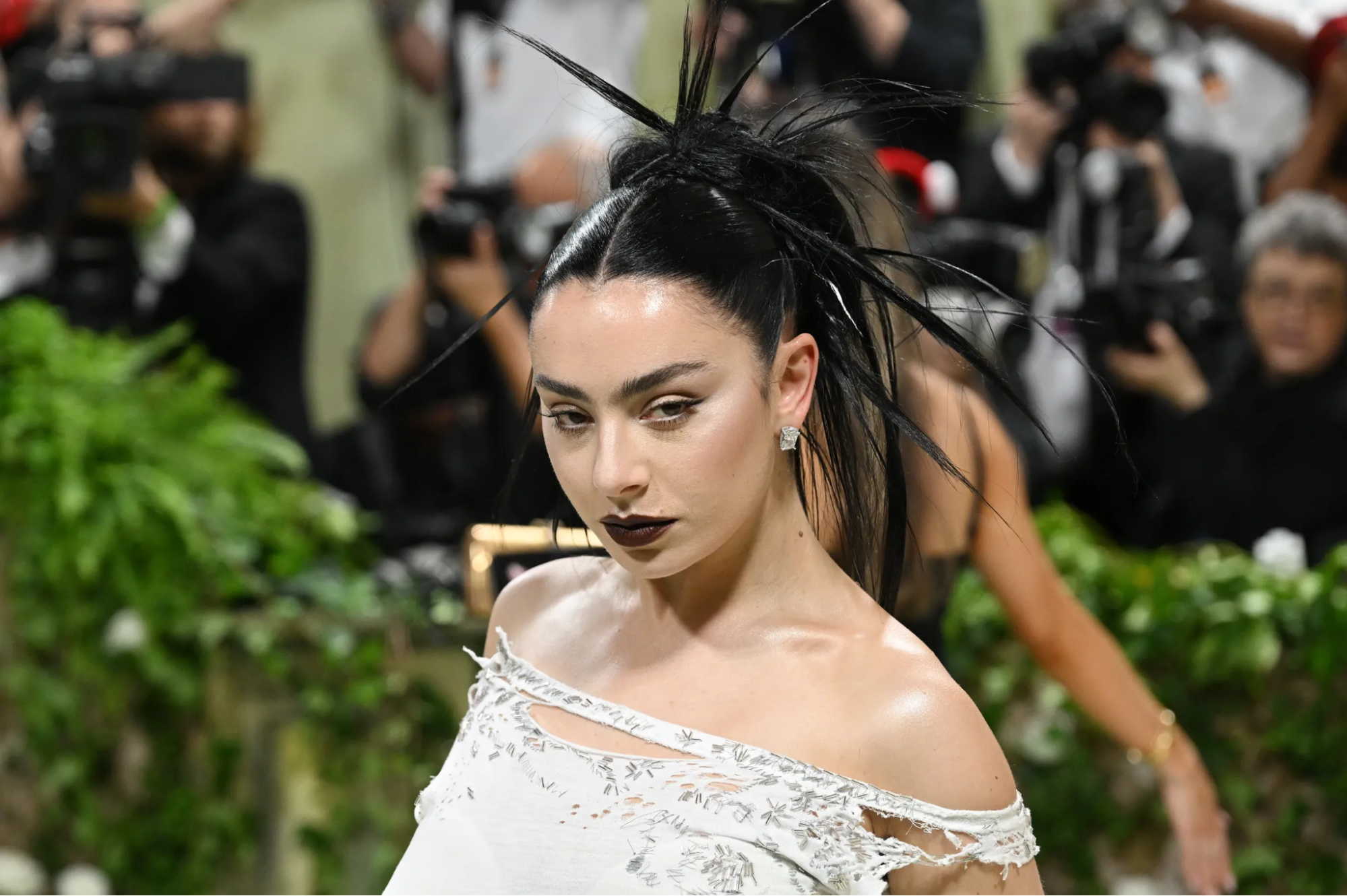 Foto da cantora Charli xcx no Met Gala. Ela é uma mulher branca com cabelos pretos. Seu penteado está com mechas espetadas para todos os lados. Seu batom é escuro assim como seu lápis de olho. Ela está olhando de canto de olho para a direita. A foto abrange seu busto até a região da cabeça, com fundo desfocado de vários fotógrafos tirando fotos. Seu vestido é branco com detalhes em cinza e retalhos como se estivesse rasgado.