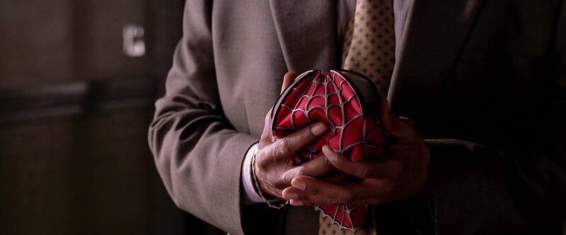 Cena do filme Homem-Aranha 2 Na imagem, o personagem Robbie segura a máscara do Homem-Aranha. Em plano detalhe, bem próximo das mãos, a imagem não mostra o rosto do personagem, apenas o peito. Ele veste um terno cinza, com gravata bege com vários losangos. Ele é um homem negro. Em seu pulso há uma pulseira de ouro. A máscara é vermelha, com lentes prateadas e teias em relevo. 